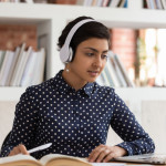 Attractive indian ethnicity girl do assignment preparing for university test or exams seated at desk in public library wearing headphones listen audio task, e-learning process educational app concept